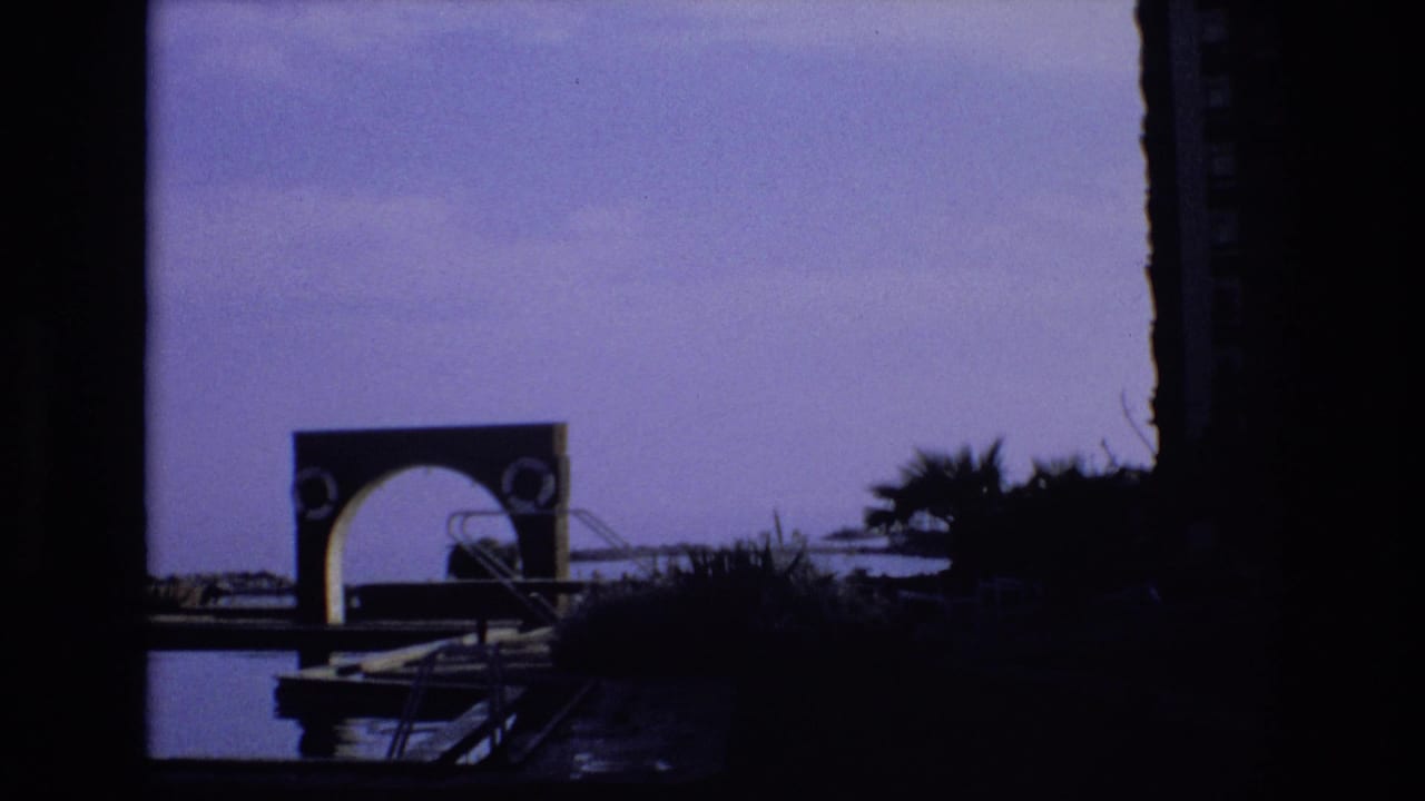 1972 Spain home-movie frame showing a dark silhouetted pool or lakeside area with an arched structure, palms, railings, and purple-tinted sky.