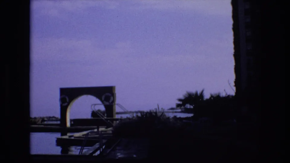 1972 Spain home-movie frame showing a dark silhouetted pool or lakeside area with an arched structure, palms, railings, and purple-tinted sky.