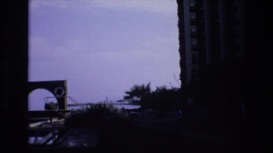 1972 Spain 8mm frame of a poolside or lakeside leisure area, with an arched architectural feature, dark palm shapes, railings, and heavy shadow.