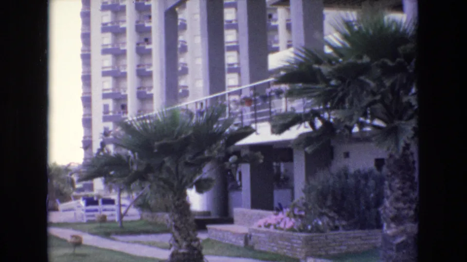 1972 Spain home-movie frame showing a residential condominium exterior with high-rise balconies, palm trees, a walkway, and planted landscaping.