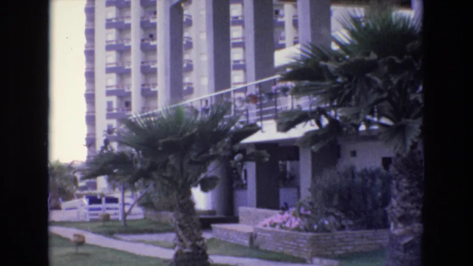 1972 Spain 8mm frame of a landscaped condominium entrance or poolside grounds with palm trees, balconies, railing, walkway, and planted flowers.