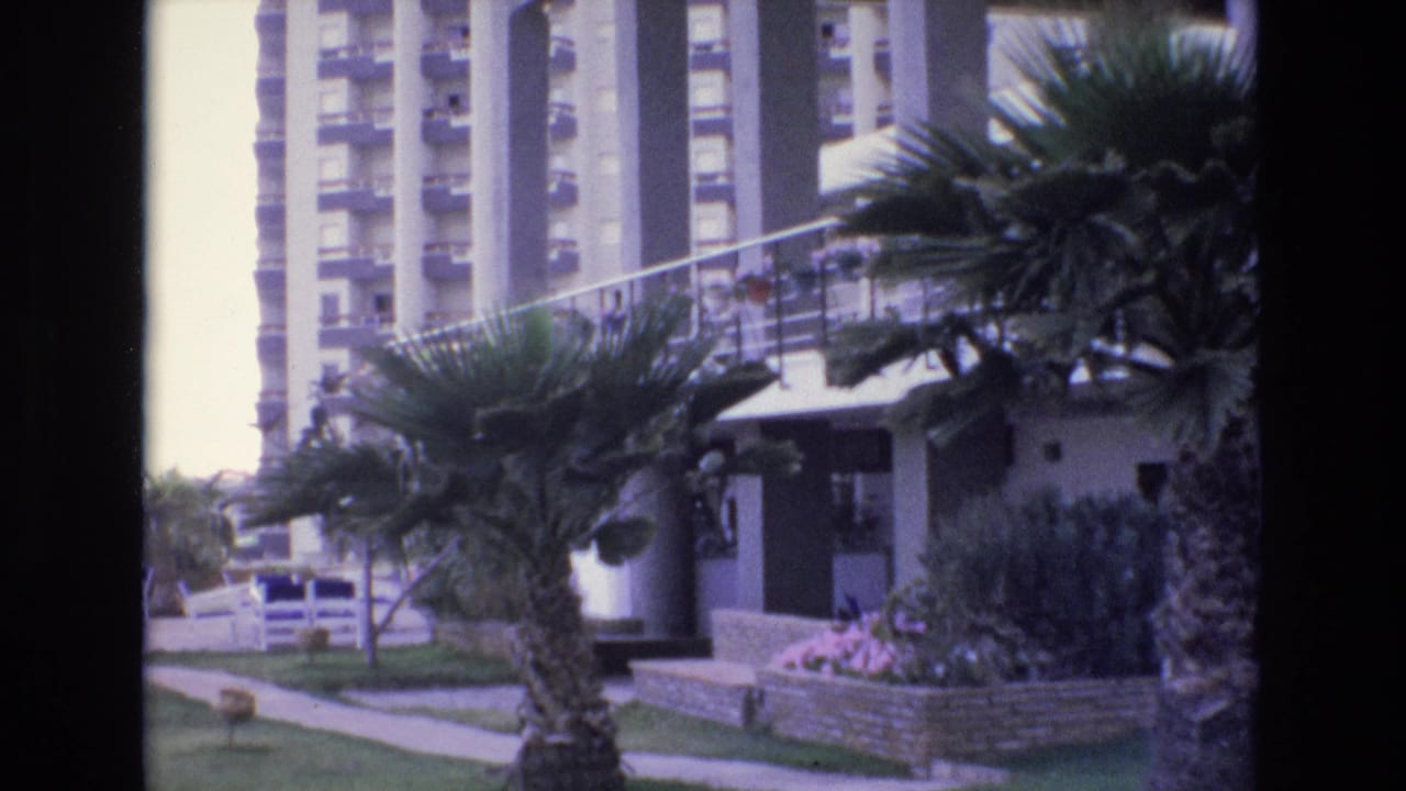 1972 Spain home-movie frame showing palm trees, a walkway, lawn, and a multi-story resort or apartment building near the camera position.