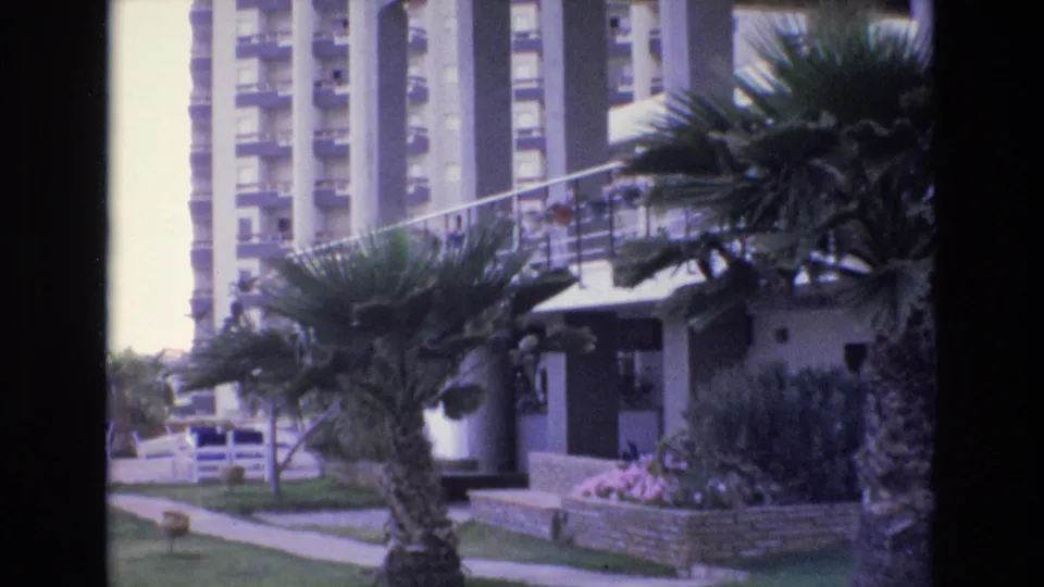 1972 Spain home-movie frame showing palm trees, a walkway, lawn, and a multi-story resort or apartment building near the camera position.