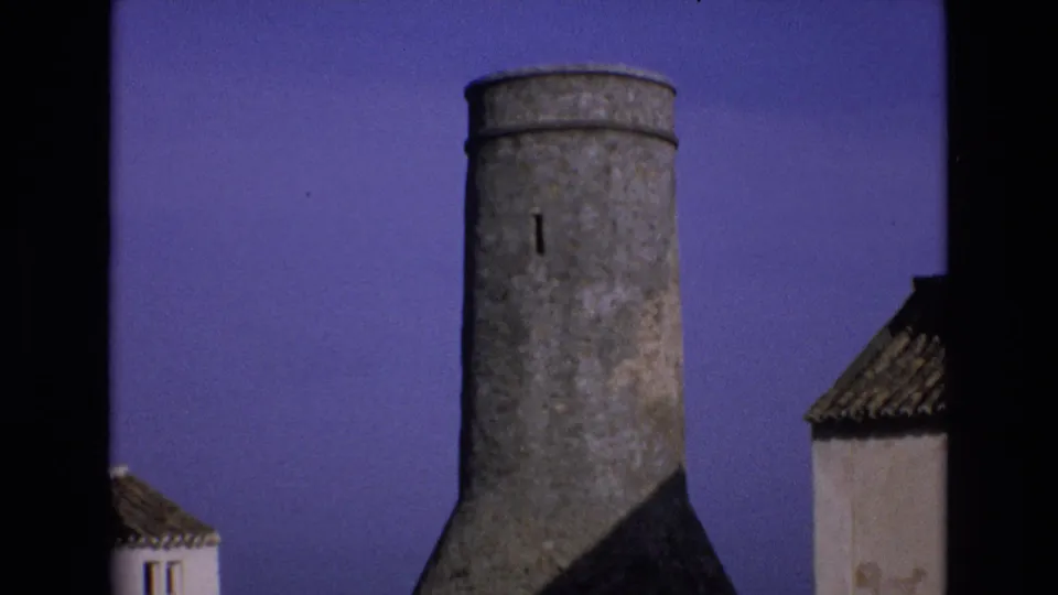 1972 Spain frame showing a weathered cylindrical stone tower with a narrow slit window, bordered by whitewashed buildings with tiled roofs.