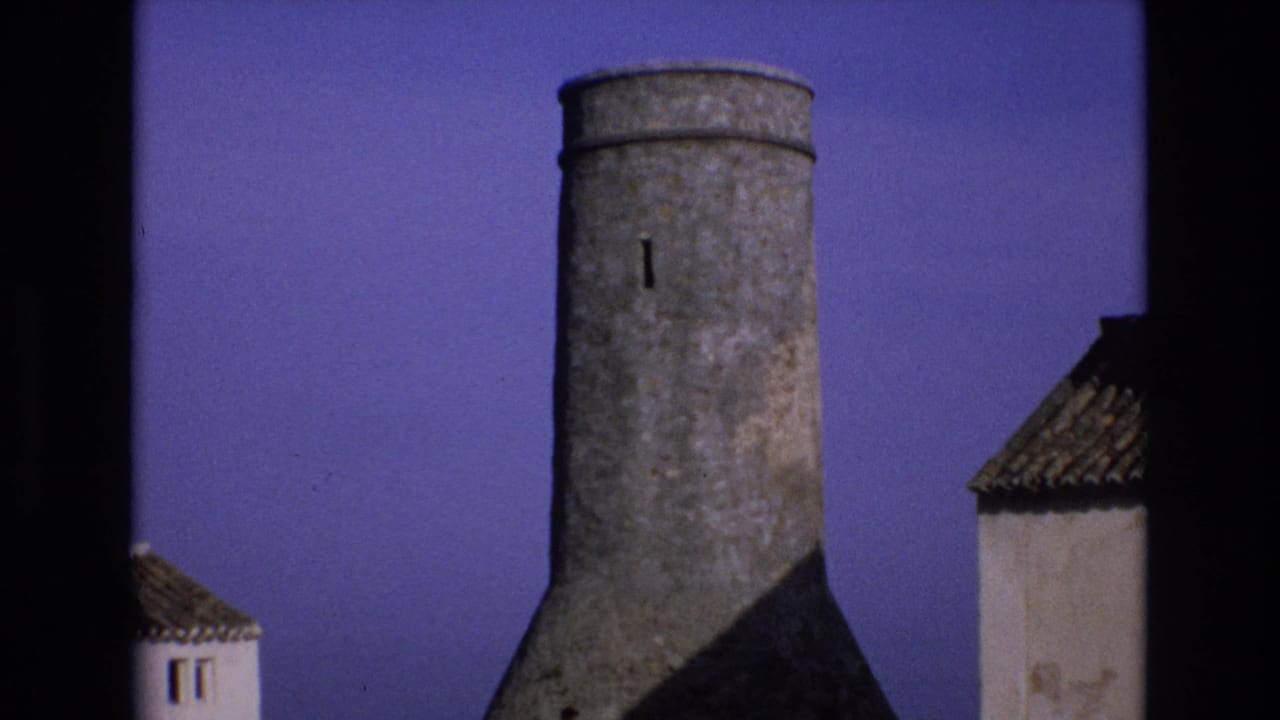 1972 Spain home-movie frame showing a weathered cylindrical stone tower with a narrow slit window between white-washed buildings and tiled roof edges.