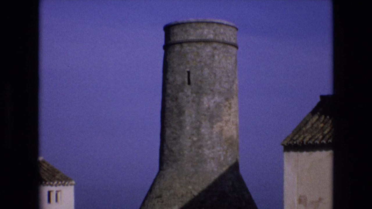 A weathered round stone tower rises above tiled roof edges in 1972 Spain, seen in soft 8mm color against a deep purple sky.