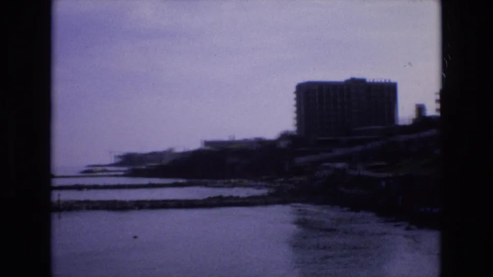 A coastal view in Spain shows calm bay water, low shoreline structures, and a prominent high-rise building in soft, dark 8mm exposure.