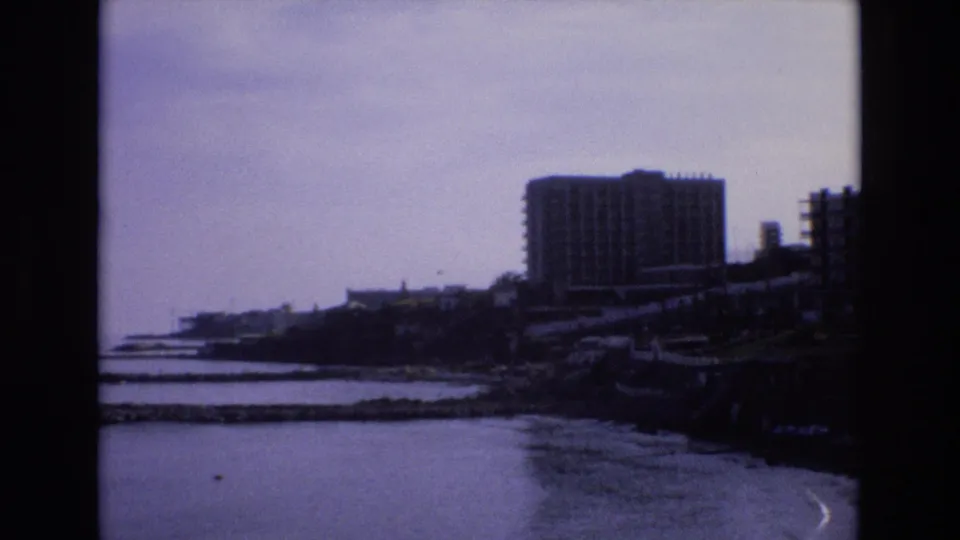 The 8mm frame looks across a Spanish bay toward a large waterfront high-rise, with breakwater lines, dark shore, and still water in the foreground.