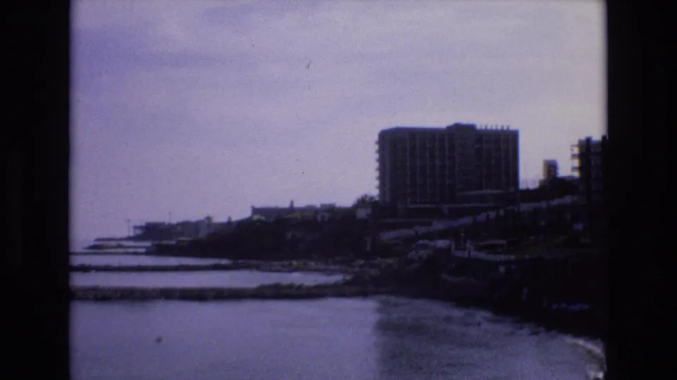A distant Spanish waterfront high-rise stands above a rocky bay edge, with still water and muted 8mm color giving the frame a subdued 1970s home-movie look.