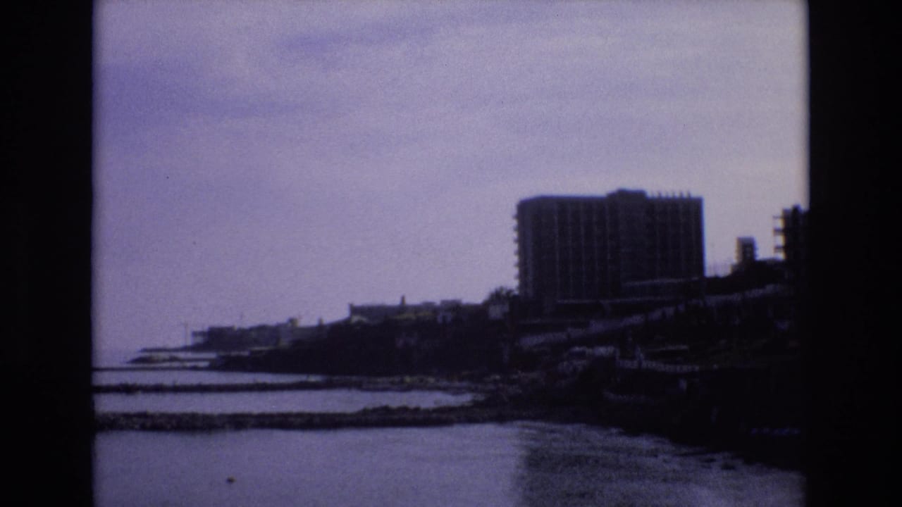 1972 Spain 8mm frame showing a distant coastal waterfront, calm water, low shoreline structures, and a large modern rectangular building in soft focus.
