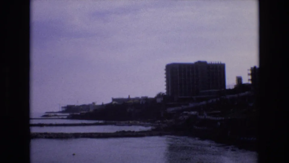 1972 Spain 8mm frame showing a distant coastal waterfront, calm water, low shoreline structures, and a large modern rectangular building in soft focus.