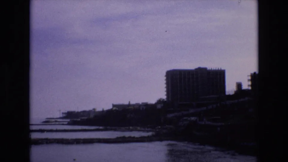 1972 Spain 8mm frame with a broad waterfront view, a prominent high-rise-like building, dark shoreline, calm water, and hazy overcast sky.