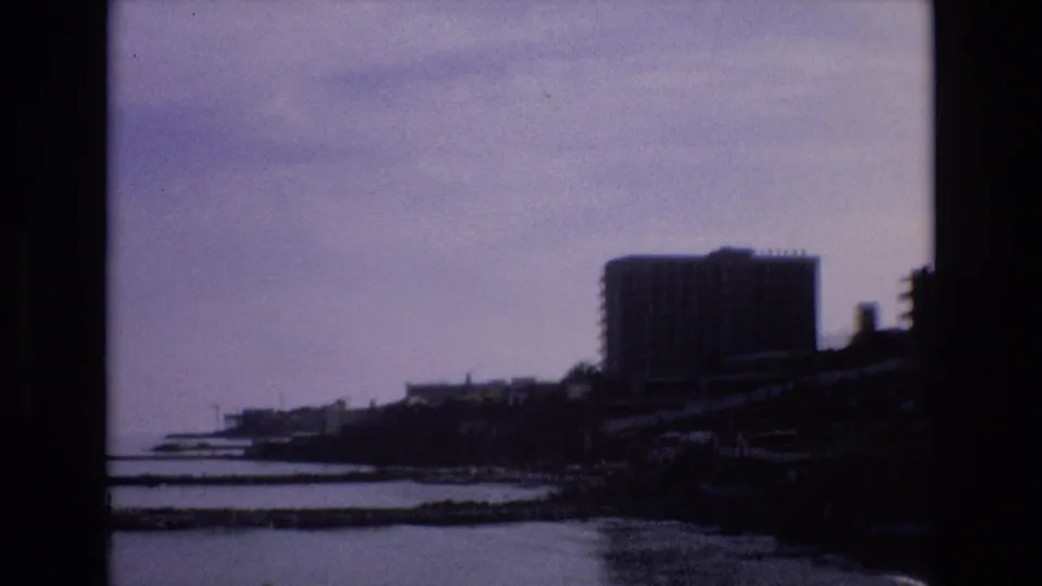 1972 Spain 8mm frame showing calm coastal water, a dark sloping shoreline, distant waterfront structures, and a large rectangular building near the horizon.