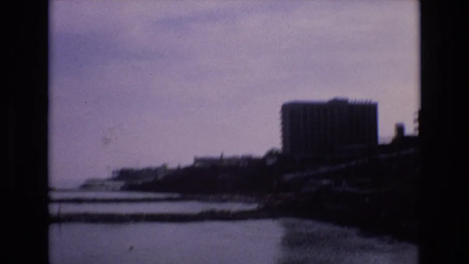 1972 Spain 8mm frame of a soft, slightly shifted waterfront view with calm water, a dark coastal edge, and a large modern building dominating the right side.