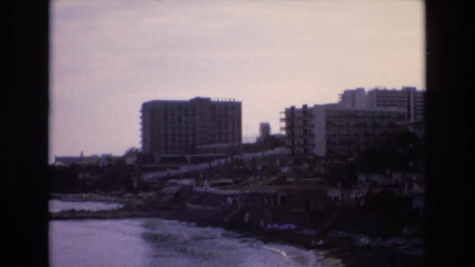 1972 Spain 8mm frame with mid-rise coastal buildings, a shoreline wall, rocks, and small waves moving along the edge of the water.