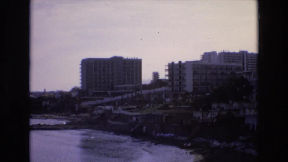 1972 Spain 8mm frame looking across calm water toward a rocky shoreline, concrete seawall, and large coastal buildings.