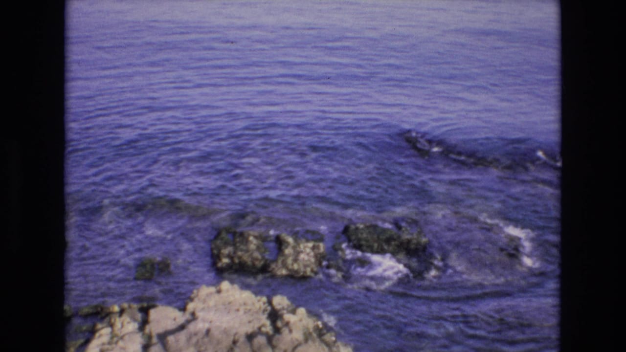 1972 Spain 8mm frame showing purple-blue coastal water rippling around dark jagged rocks beside a pale rocky edge.