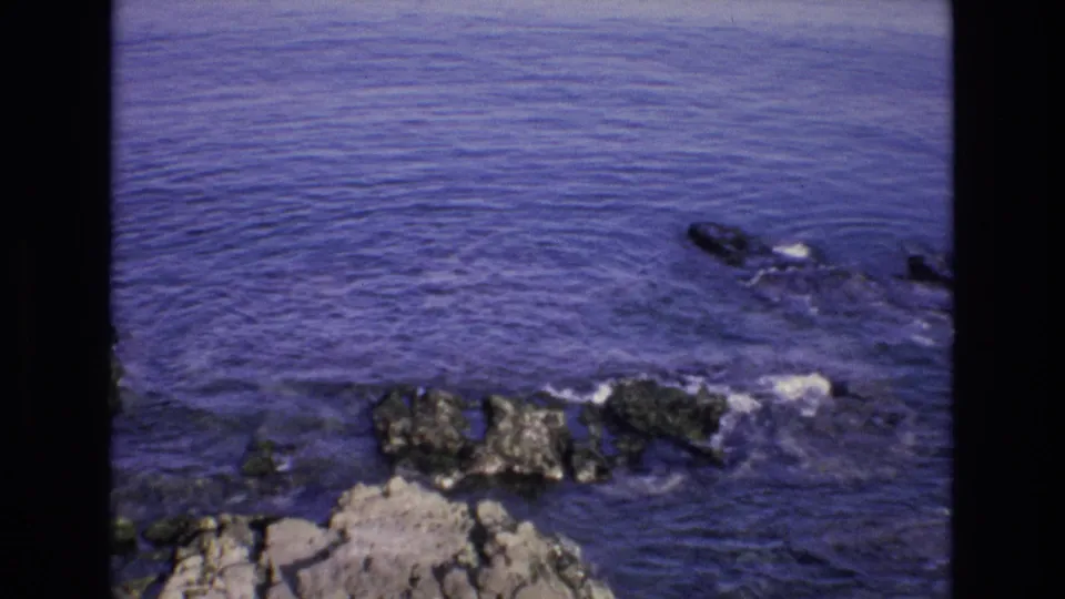 1972 Spain 8mm frame showing a small wave and foamy wash approaching jagged shoreline rocks in saturated blue-purple water.