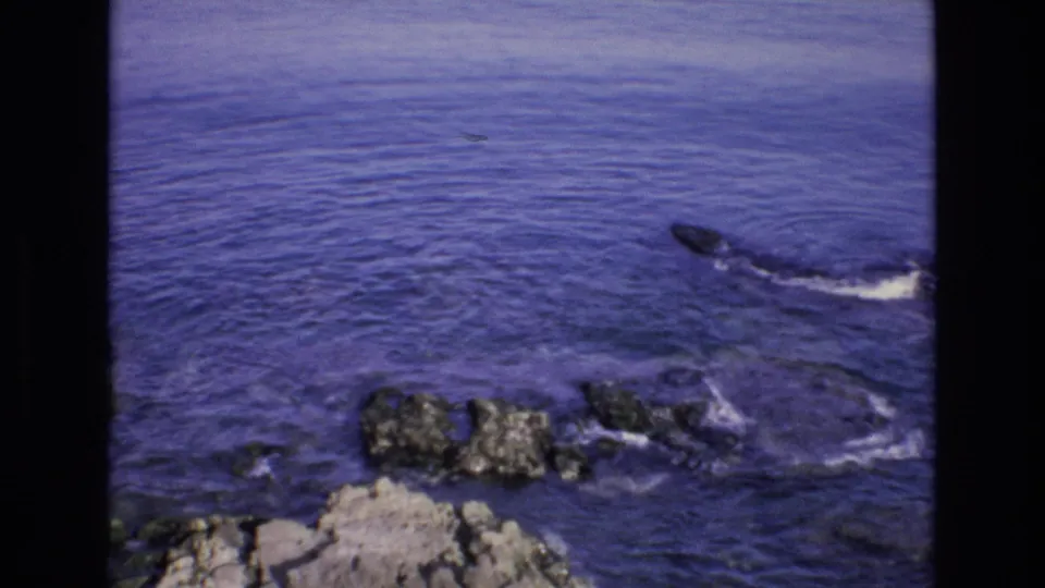 1972 Spain 8mm frame showing foamy surf swirling around dark shoreline rocks, with soft grain and black frame edges visible.
