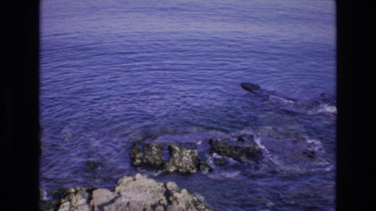 1972 Spain 8mm frame showing a blue coastal sea with scattered rocks near the shoreline, bordered by the dark edges of the film scan.