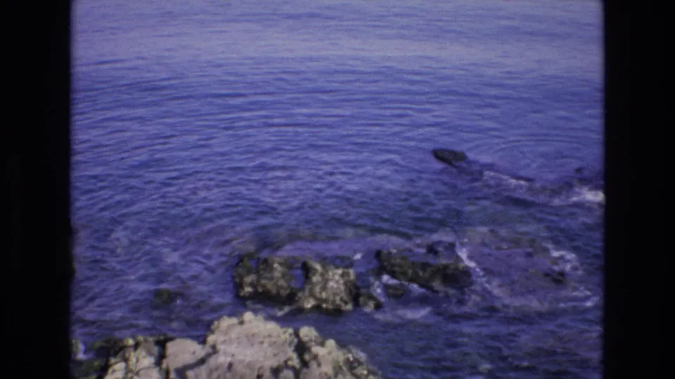 1972 Spain 8mm frame showing a blue coastal sea with scattered rocks near the shoreline, bordered by the dark edges of the film scan.