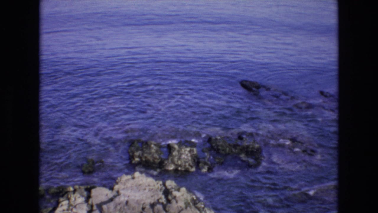 Rocky shoreline and blue water with a dark vertical film border through the frame.