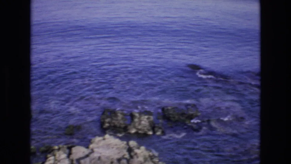 1972 Spain 8mm frame looking down toward a rocky shoreline and rippled blue sea, with grain and saturated color typical of home-movie footage.