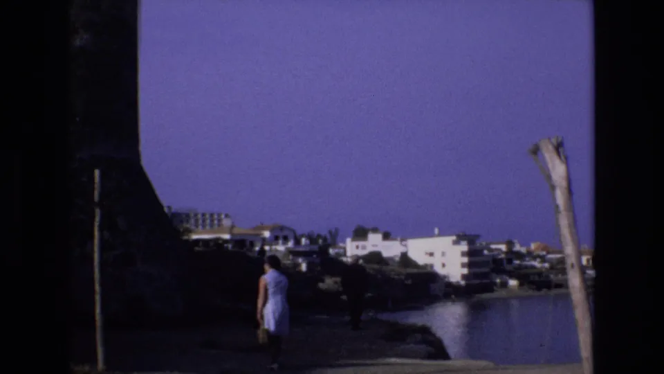 1972 Spain 8mm frame showing a woman in a light dress walking on a waterfront path toward white coastal buildings under a clear blue sky.