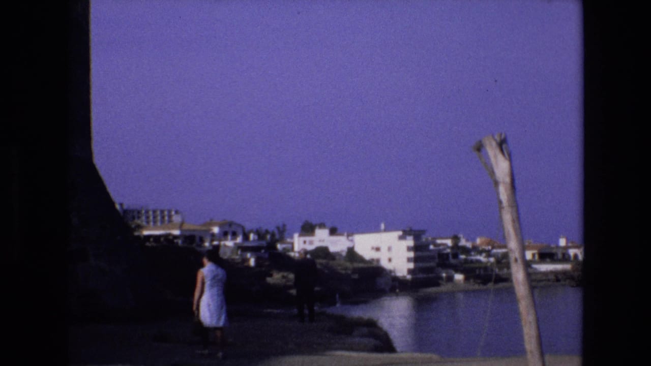 1972 Spain 8mm frame showing a woman in a light dress near a rocky shoreline, calm cove water, white coastal buildings, and a weathered pole under a purple-blue sky.