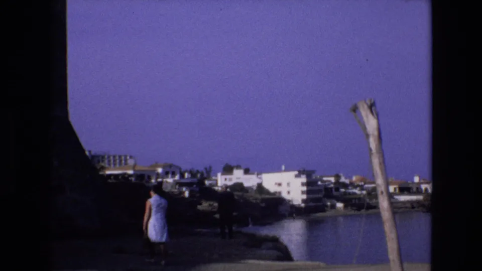1972 Spain 8mm frame showing a woman in a light dress near a rocky shoreline, calm cove water, white coastal buildings, and a weathered pole under a purple-blue sky.