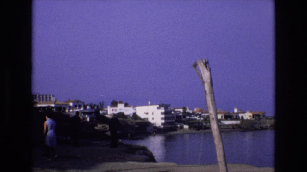 Woman walking farther along the shaded shoreline with white buildings across the cove.
