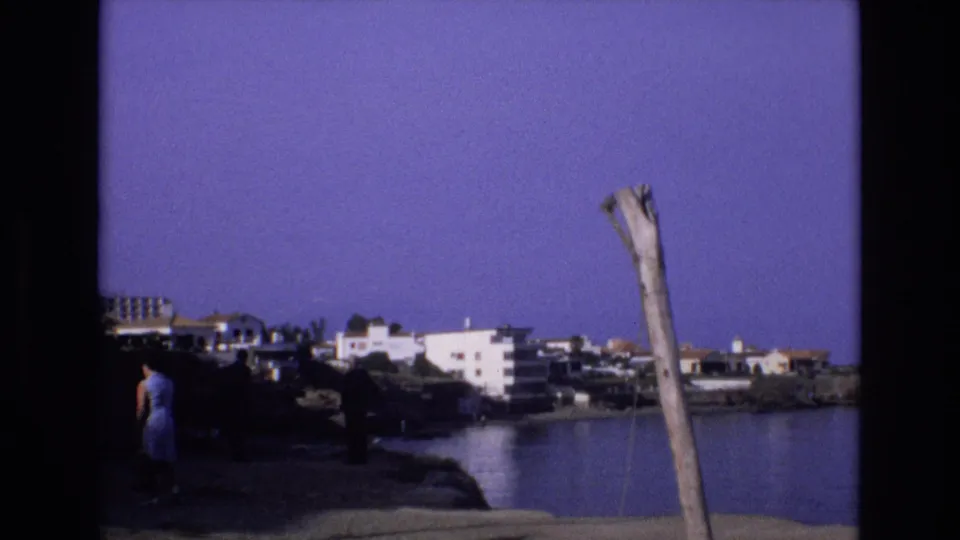1972 Spain 8mm frame with a woman moving along the dark rocky edge of a cove, backed by white hillside buildings and a prominent weathered pole in the foreground.