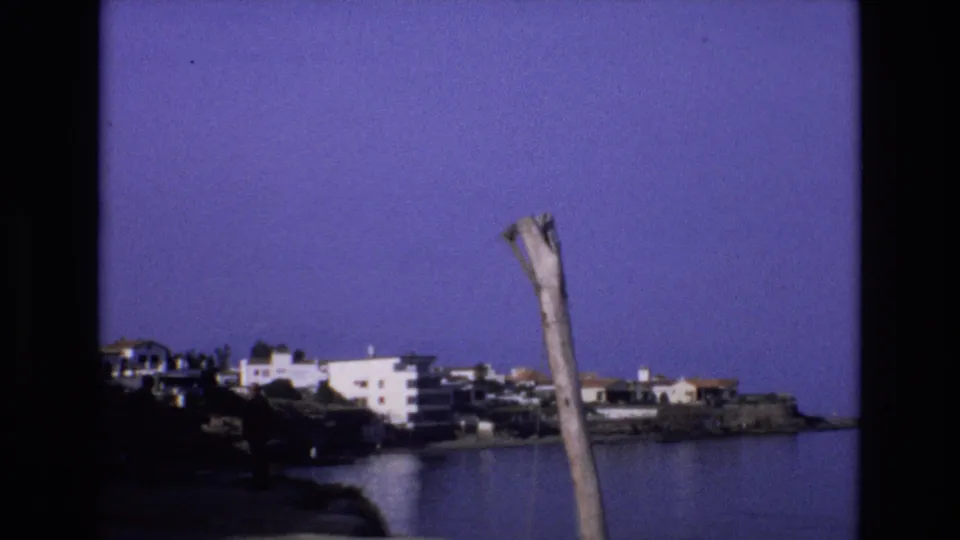 1972 Spain 8mm frame looking across a quiet cove toward white coastal buildings, with a distant person on the rocky shore and a weathered wooden pole cutting into the sky.
