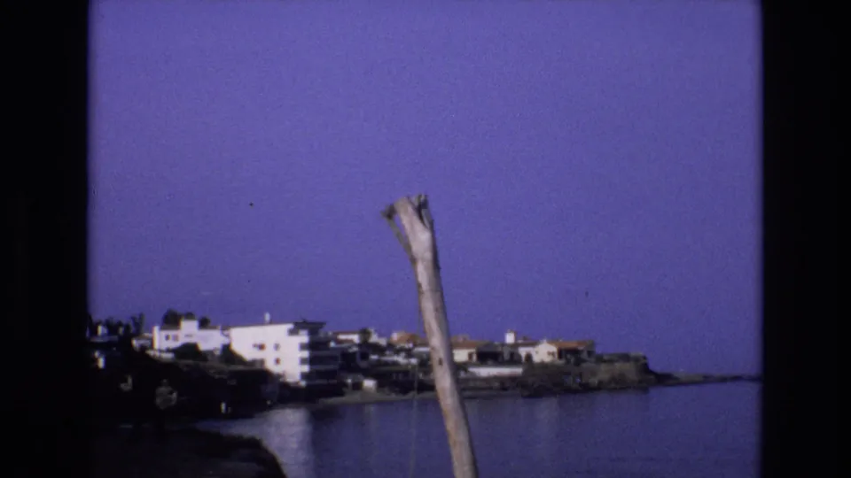 1972 Spain 8mm frame of calm cove water and white coastal buildings on the opposite shore, framed by a weathered wooden pole and dark foreground vegetation.