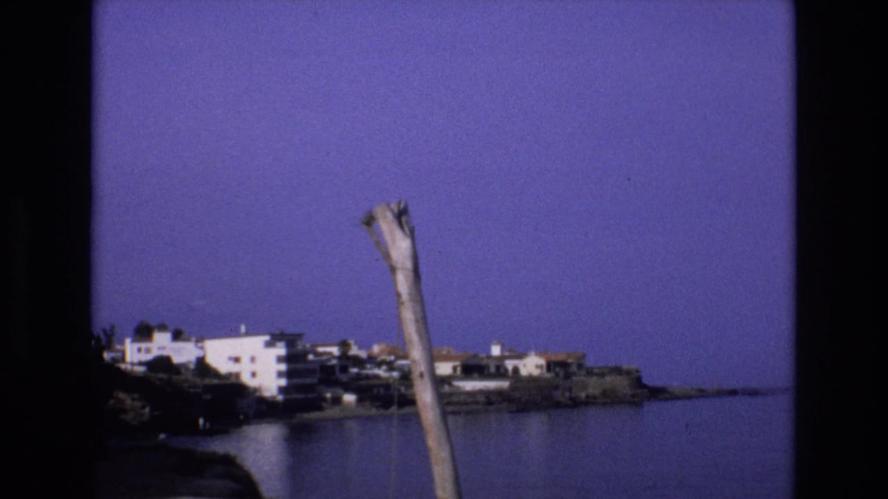 1972 Spain 8mm frame showing a calm coastal bay, white shoreline buildings, rocky waterfront, deep blue-purple sky, and a weathered pole in the foreground.