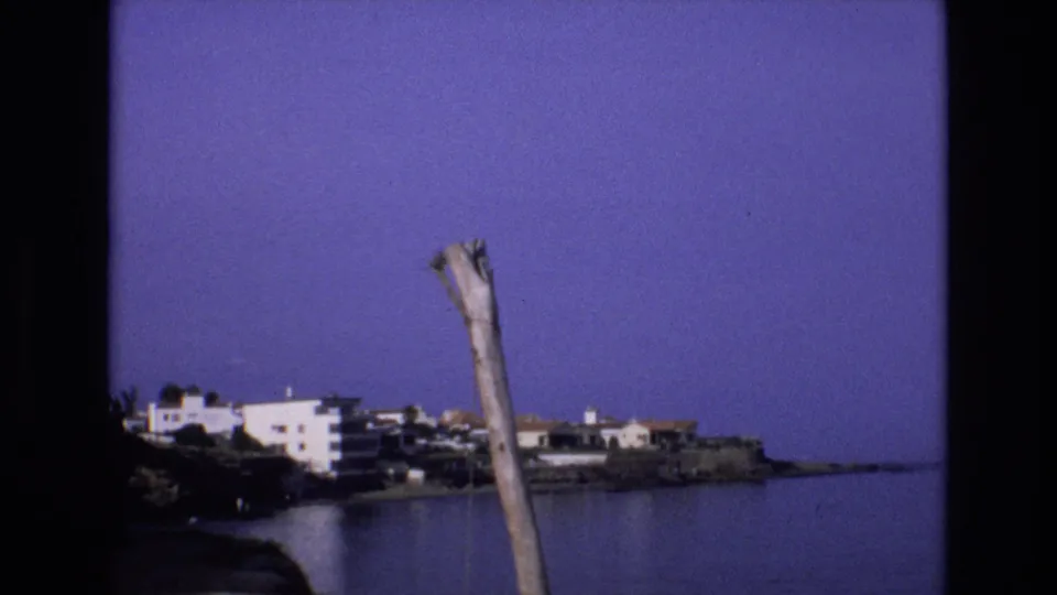 1972 Spain 8mm frame showing a calm coastal bay, white shoreline buildings, rocky waterfront, deep blue-purple sky, and a weathered pole in the foreground.