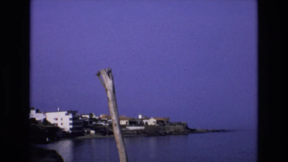 1972 Spain 8mm frame of a coastal town across calm water, with white buildings on a rocky point and a weathered pole crossing the foreground.