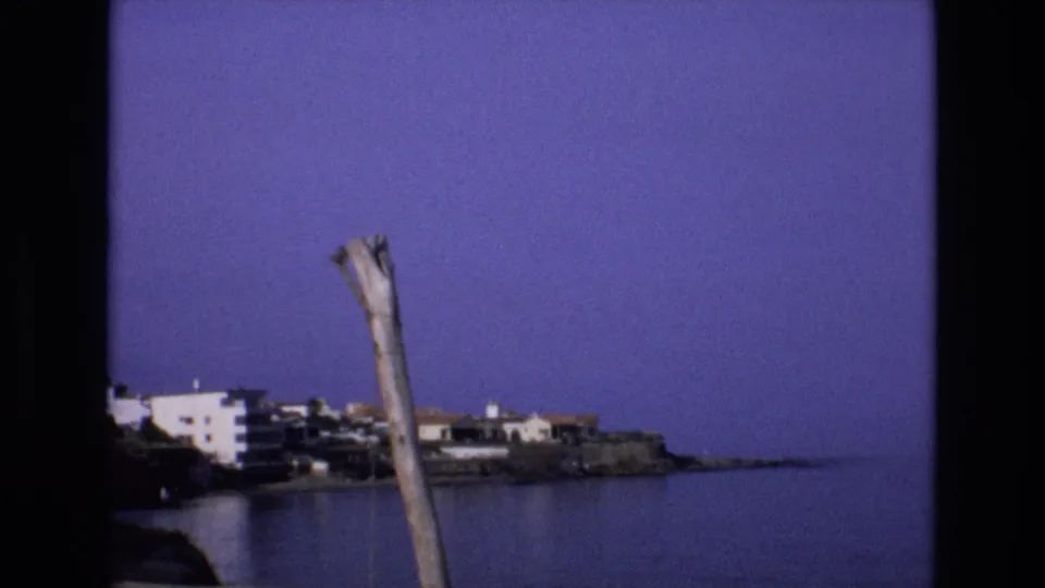 1972 Spain 8mm frame holding on a quiet bay scene, with a weathered wooden pole, white waterfront buildings, rocky shoreline, and open water.