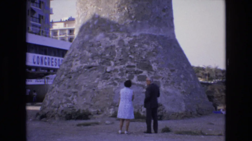 1972 Spain 8mm frame showing two adult visitors admiring a massive stone tower or monument, with modern buildings and a partial "CONGRESO" sign in the background.