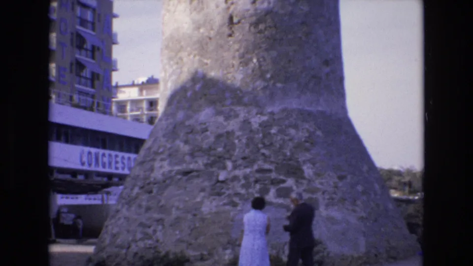 The handheld 8mm frame centers the broad weathered stone tower base in Spain, with the adult couple partly visible near the bottom and modern buildings along the left edge.