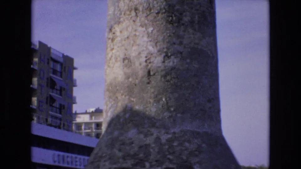 1972 Spain 8mm frame with the rough surface of a concrete tower dominating the foreground and hotel or congress-area buildings visible behind it.