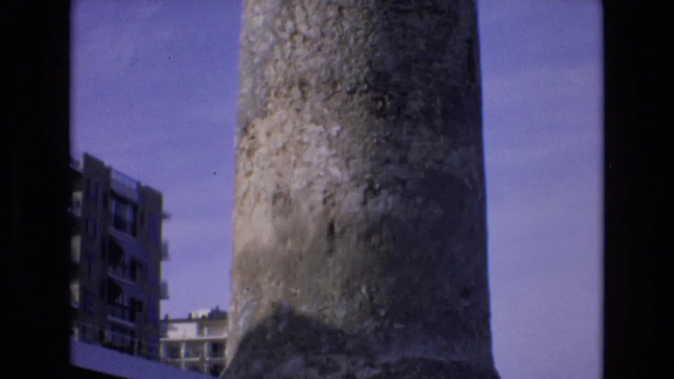 1972 Spain home-movie frame showing more of the vertical concrete tower, with a multi-story building and partial signage at the left edge.