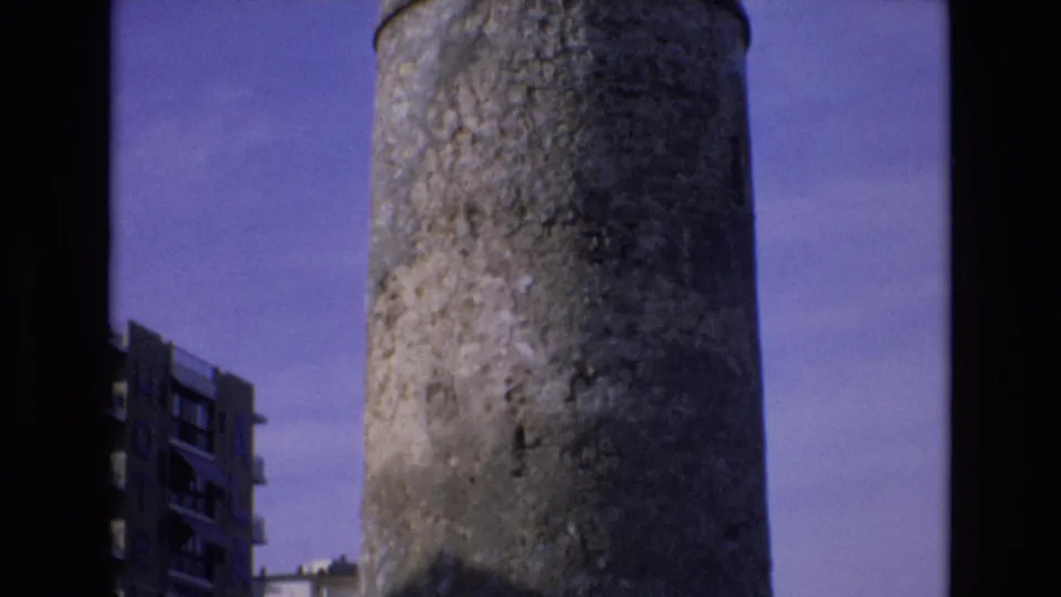 1972 Spain 8mm frame holding on the upper section of a weathered concrete tower, with a hotel-like building partly visible at the left edge.