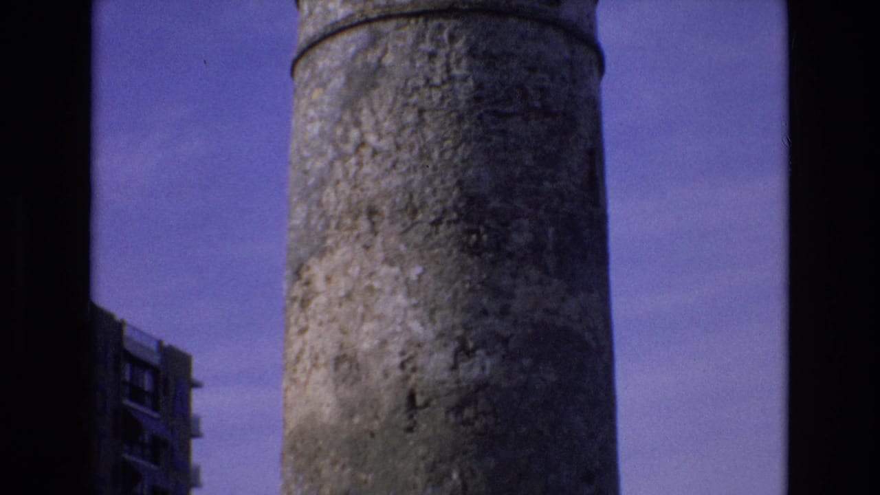 1972 Spain 8mm frame showing a low-angle view of a weathered cylindrical concrete tower, with a partial modern building at left and dark film borders around the image.