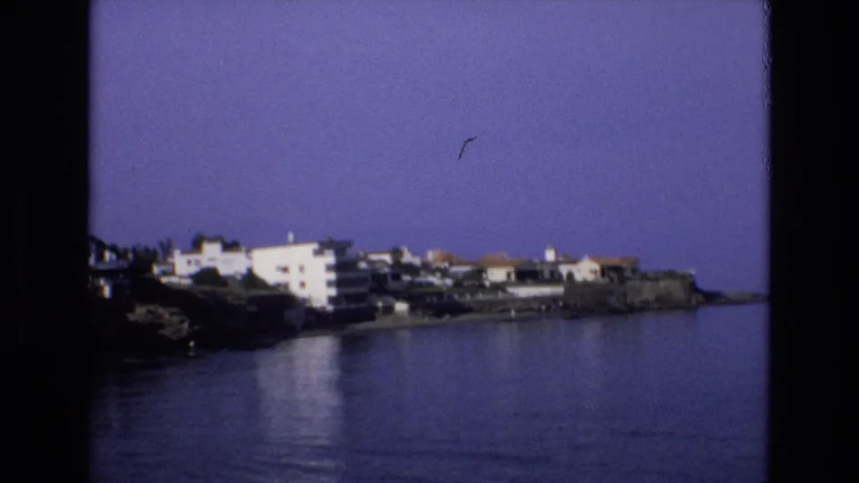 1972 Spain 8mm frame showing white waterfront houses with terracotta-toned roofs along a rocky shoreline, viewed across calm water under a hazy purple-blue sky.