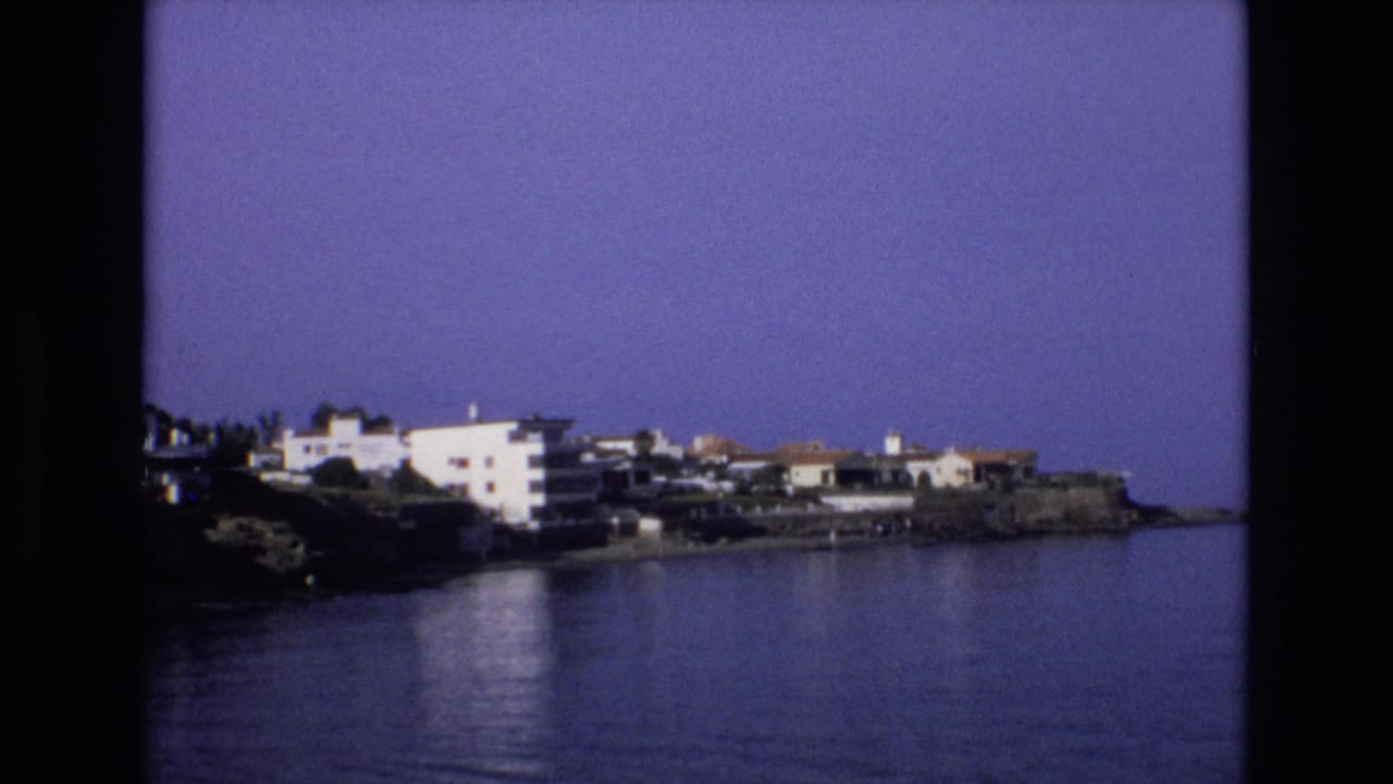 1972 Spain home-movie frame showing white apartments and houses along a rocky coast with calm blue water in the foreground.