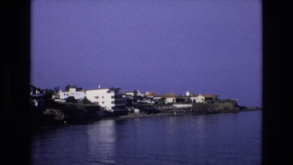 1972 Spain home-movie frame showing white apartments and houses along a rocky coast with calm blue water in the foreground.