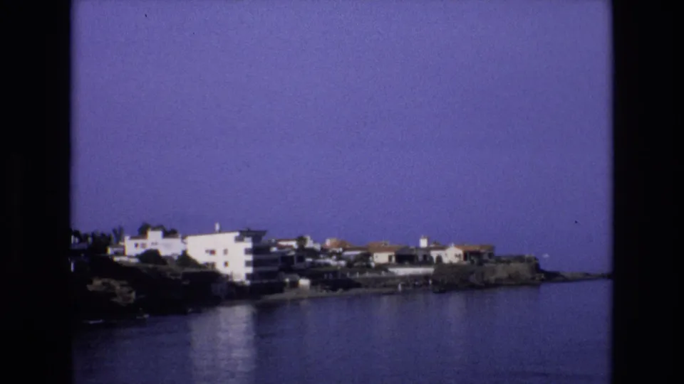 A steady 8mm frame from Spain in 1972 shows white shoreline buildings, dark rocks, calm water, and a broad blue sky.