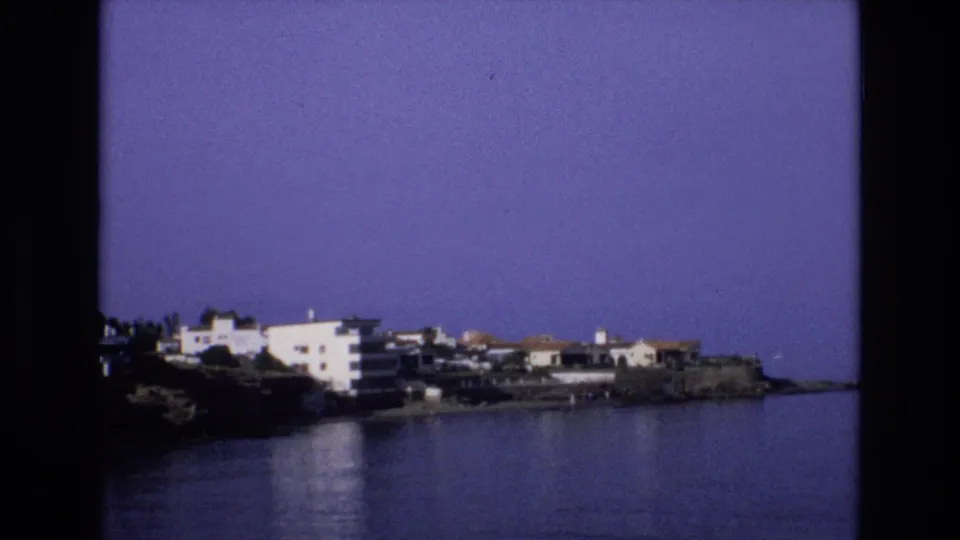 Final 1972 Spain frame with white seaside buildings on a rocky edge, calm blue water, faint reflections, and visible 8mm frame borders.