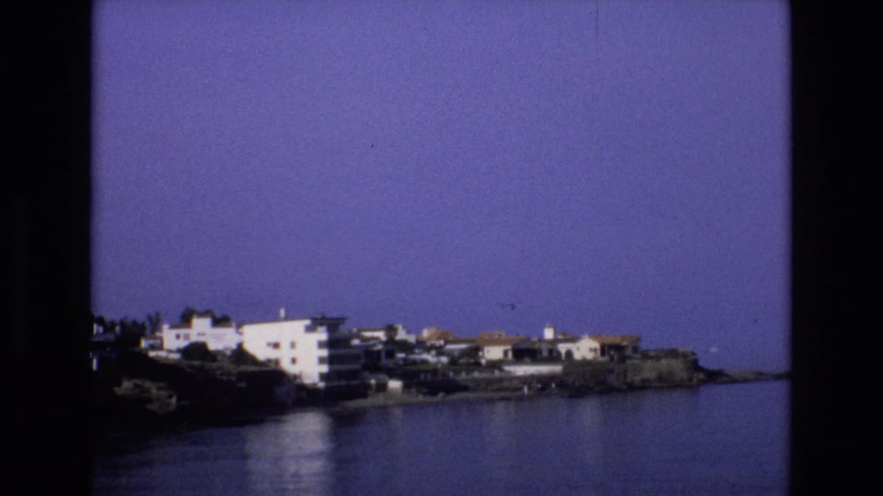 1972 Spain home-movie frame showing white and cream buildings along a rocky shoreline with calm water in the foreground and a clear blue-purple sky above.